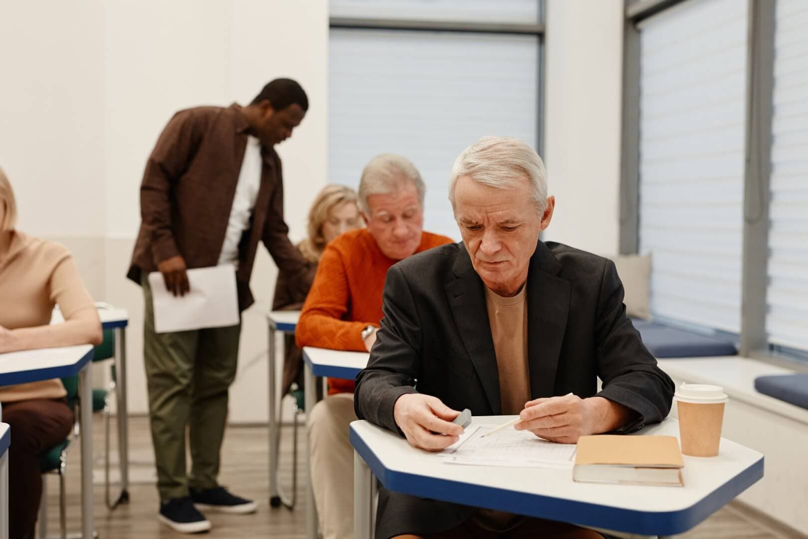 Elderly students taking an exam in a classroom, with a teacher supervising and a book and coffee cup on a desk. - Home Instead Poole