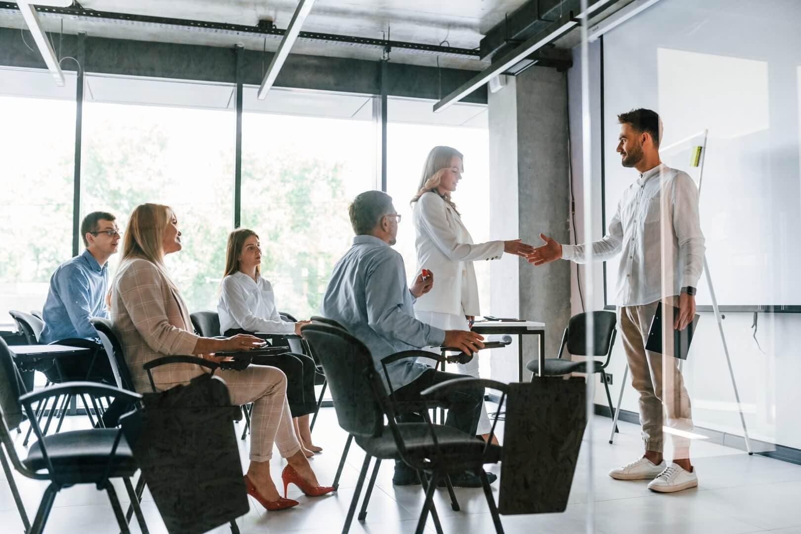 A group of people in a modern office setting, with two individuals shaking hands in front of a presentation screen. - Home Instead