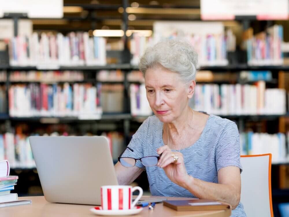 Elderly woman working on a laptop in a library, adjusting her glasses, with a coffee cup and notebook on the table. - Home Instead Southampton