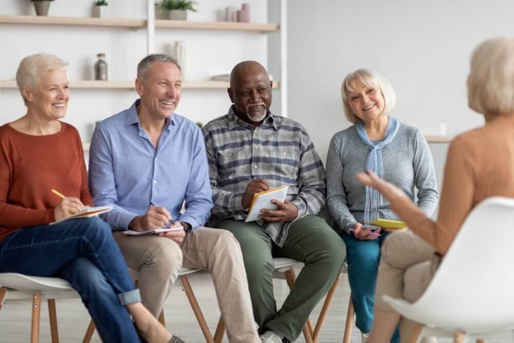 A group of four older adults sits in chairs, attentively engaging in a discussion with a fifth person who is standing. - Home Instead