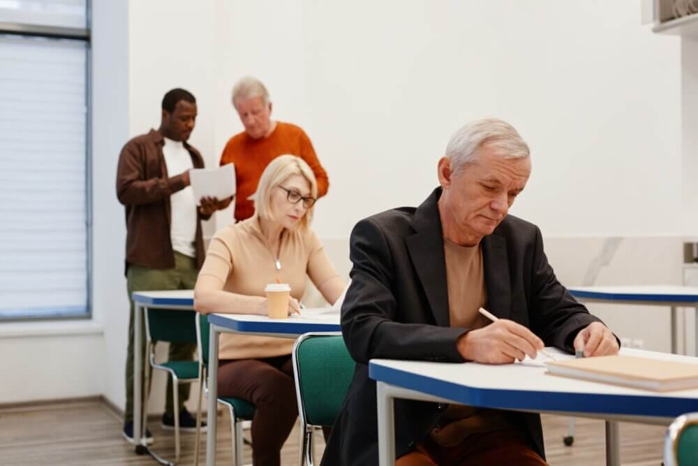 Several people of varying ages are taking a written test in a classroom setting. - Home Instead