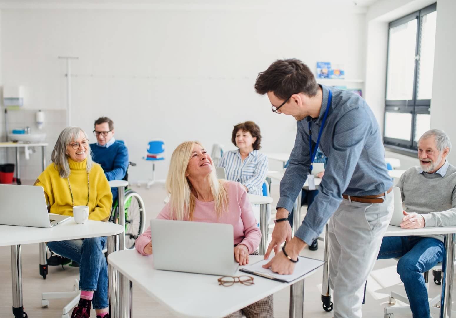 An instructor helps a smiling senior woman in a classroom with other seniors, some with laptops, in a bright, airy room. - Home Instead