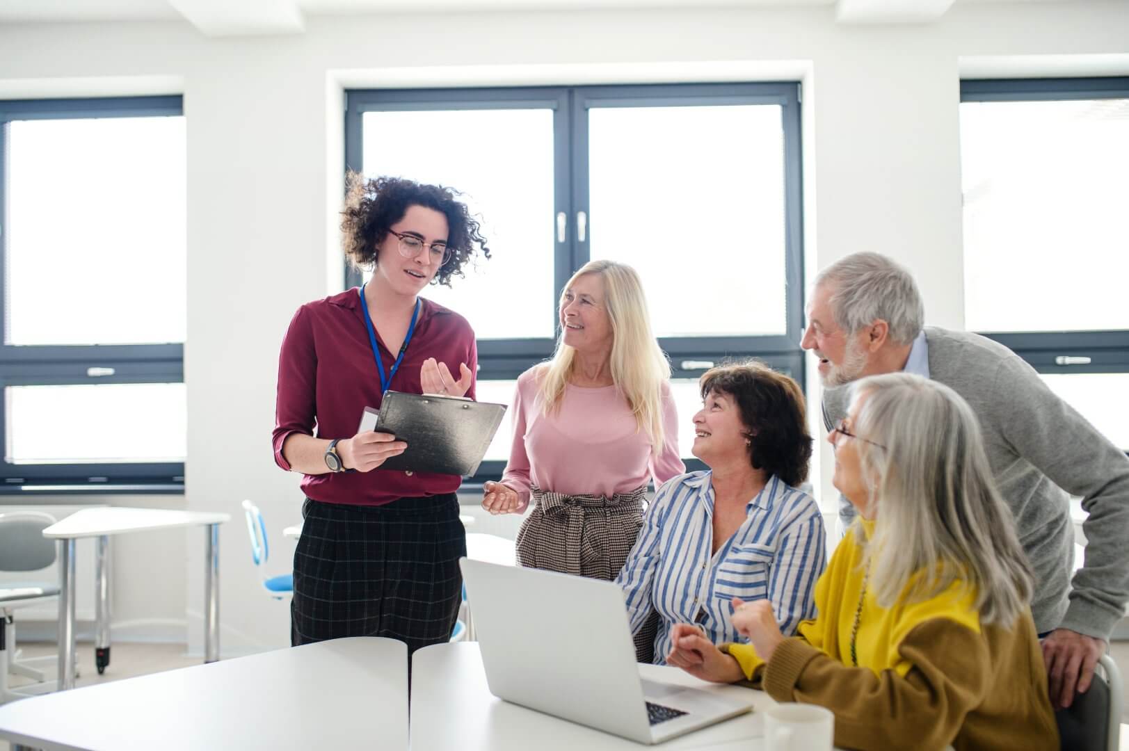 A person presents information on a tablet to a group of four older adults gathered around a table in a well-lit room. - Home Instead
