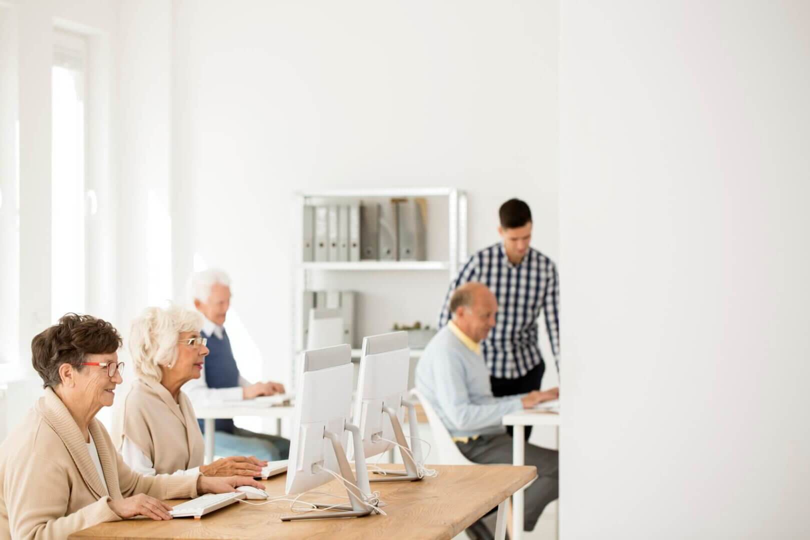 Elderly individuals in front of computers, learning with assistance from a young man in a bright, modern classroom. - Home Instead Southampton