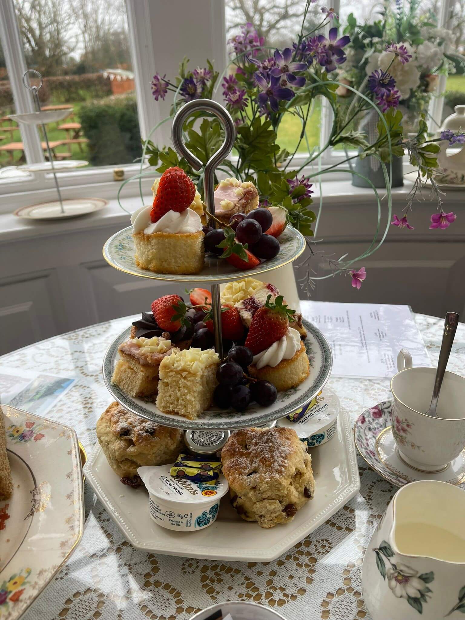 A three-tiered cake stand with assorted desserts, scones, and fruits on a table with a lace cloth near a window with flowers. - Home Instead
