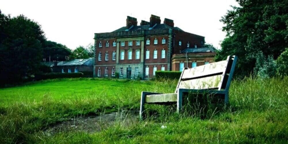 An old brick building stands in the background of a grassy field with a wooden bench in the foreground. - Home Instead