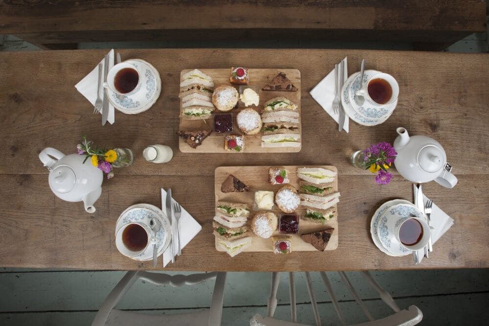 A wooden table with a tea set, floral arrangements, sandwiches, pastries, and cups of tea, viewed from above. - Home Instead