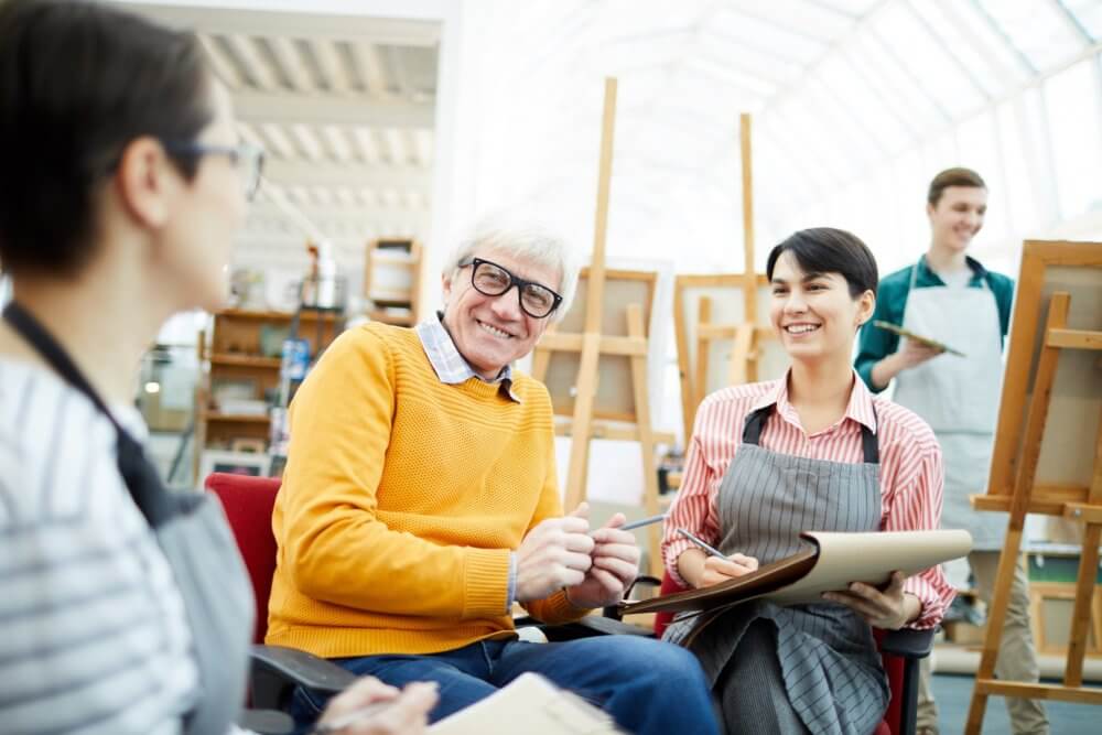A group of people in an art class, smiling and engaging in conversation, holding sketchbooks with easels in the background. - Home Instead