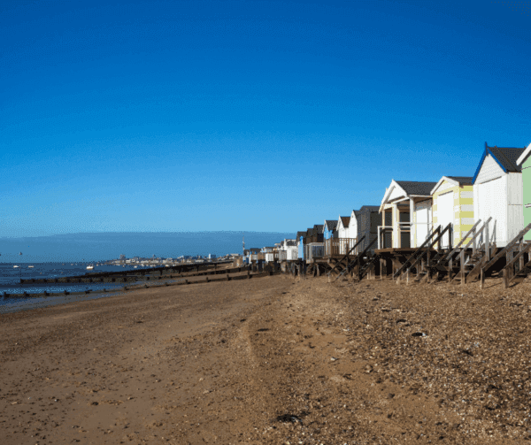 Row of colorful beach huts on a sandy shore under a clear blue sky, with the sea and distant harbor on the left. - Home Instead