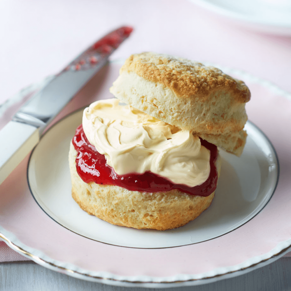 A scone topped with clotted cream and strawberry jam on a plate, with a knife smeared with jam beside it. - Home Instead