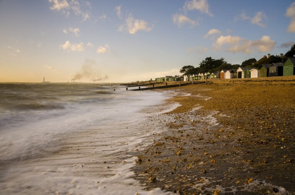 A pebbly beach with waves, colorful beach huts, and distant industrial buildings under a cloudy sky at sunset. - Home Instead