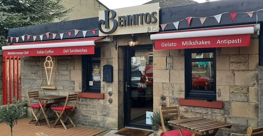 A small deli with red awnings, outdoor seating, and signs for Mediterranean deli, Italian coffee, deli sandwiches, and gelato. - Home Instead