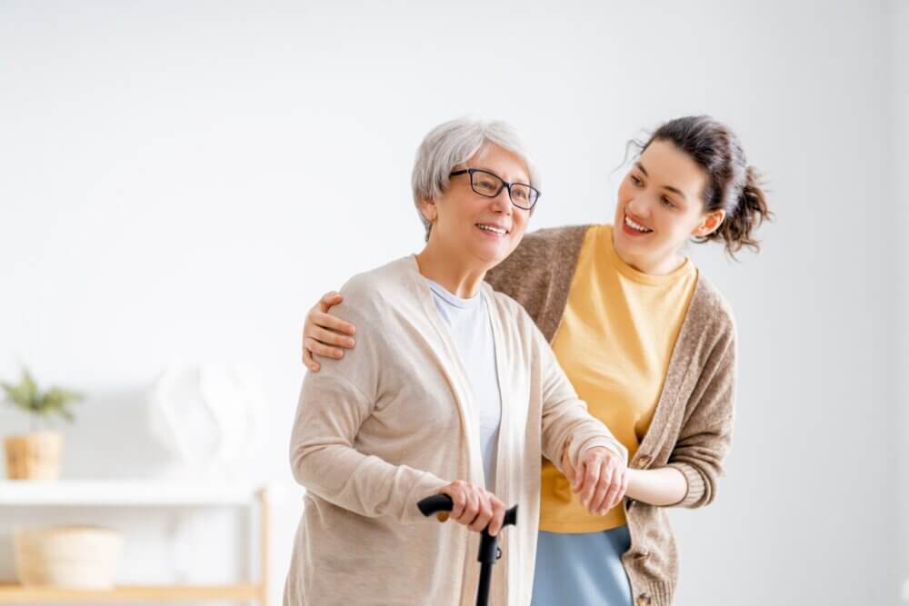 A young woman supports an elderly woman with a cane, both smiling in a bright, airy living room. - Home Instead