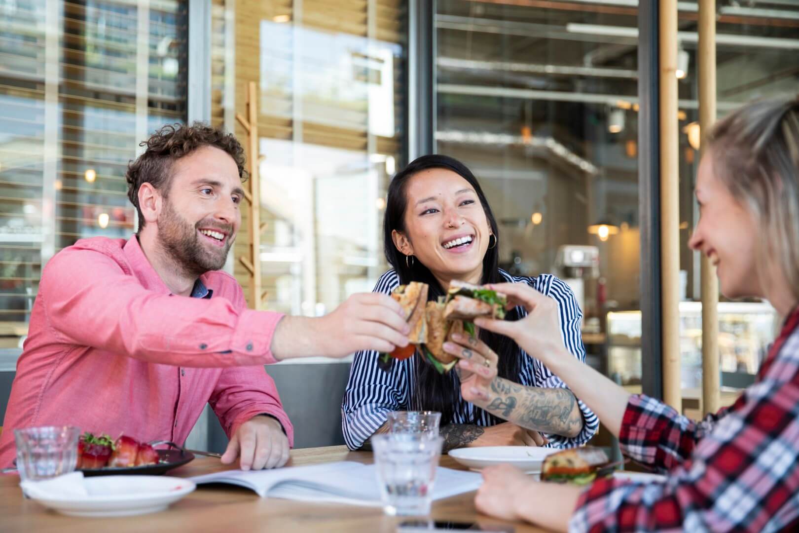 Three friends sitting at a restaurant table, laughing and clinking their sandwiches together. - Home Instead