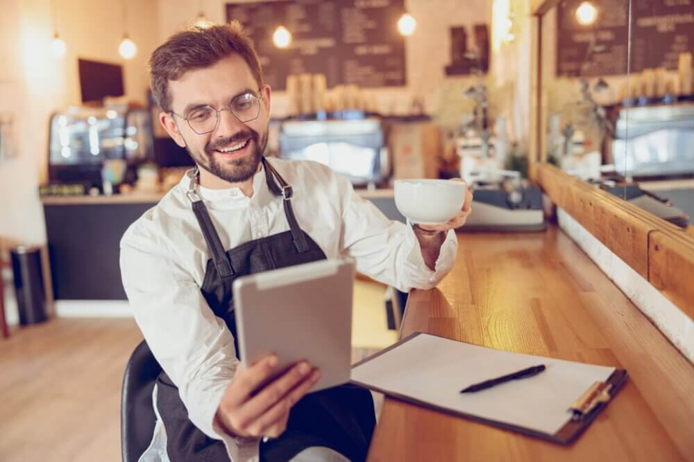 Smiling barista with glasses holds a cup of coffee and uses a tablet at a wooden counter in a cozy café. - Home Instead