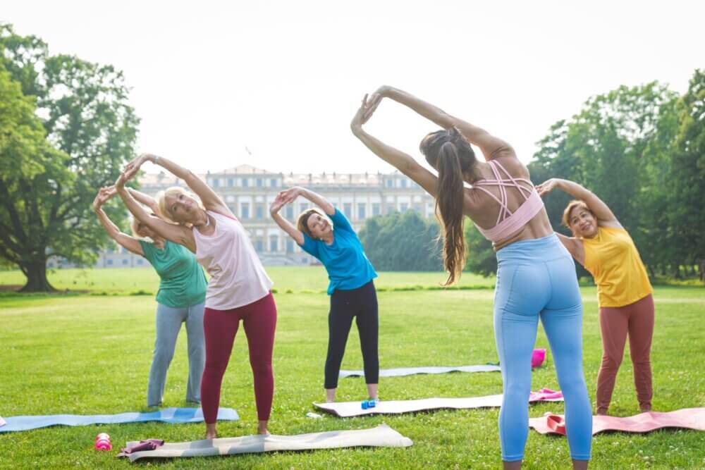 A group of women stretching together on yoga mats in a park, with a large building in the background. - Home Instead