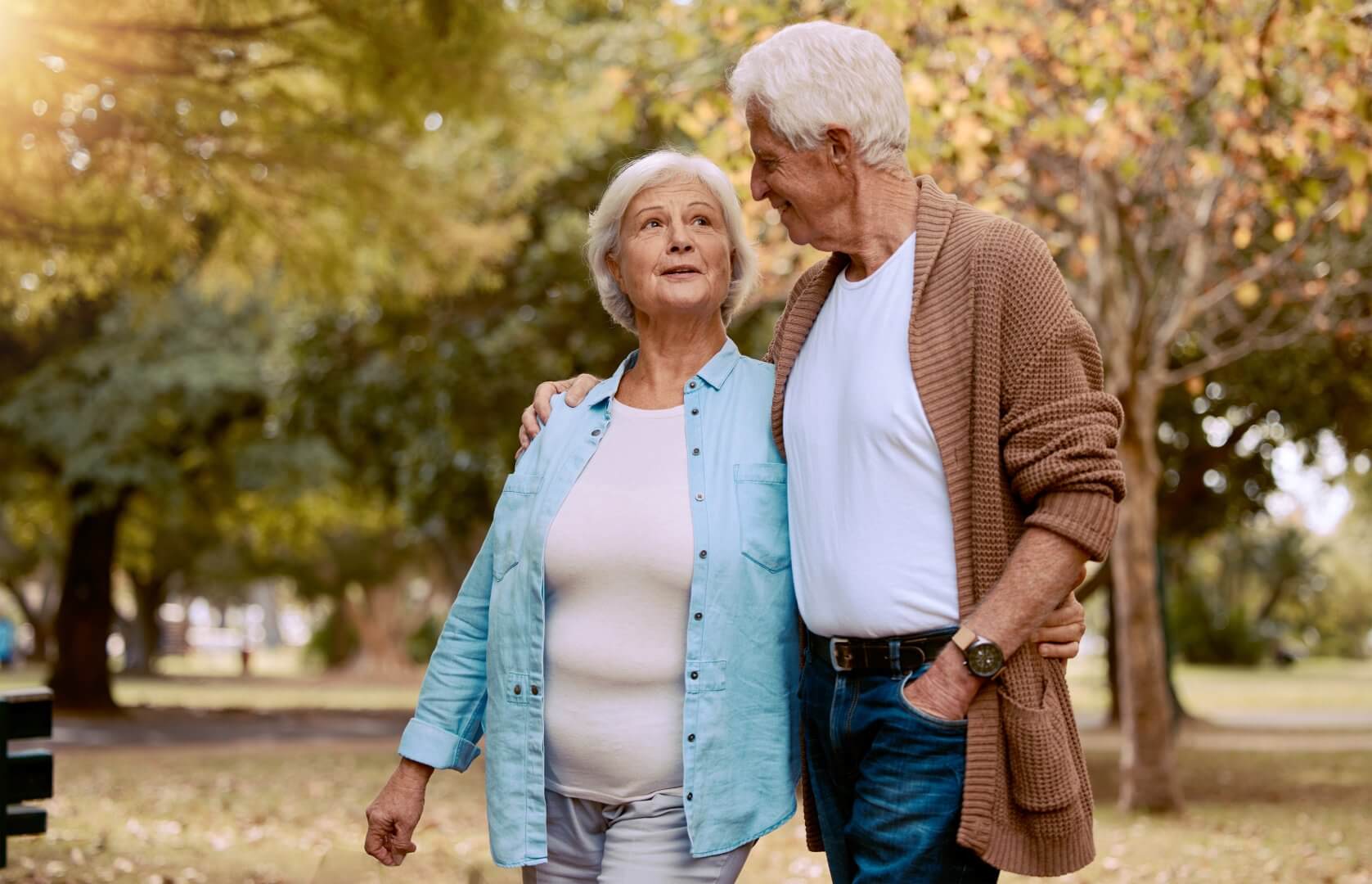Elderly couple walking arm in arm through a park with trees and fall leaves in the background. - Home Instead