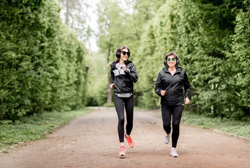 Two people jogging on a forest path, wearing sunglasses, headphones, and black athletic wear, surrounded by greenery. - Home Instead
