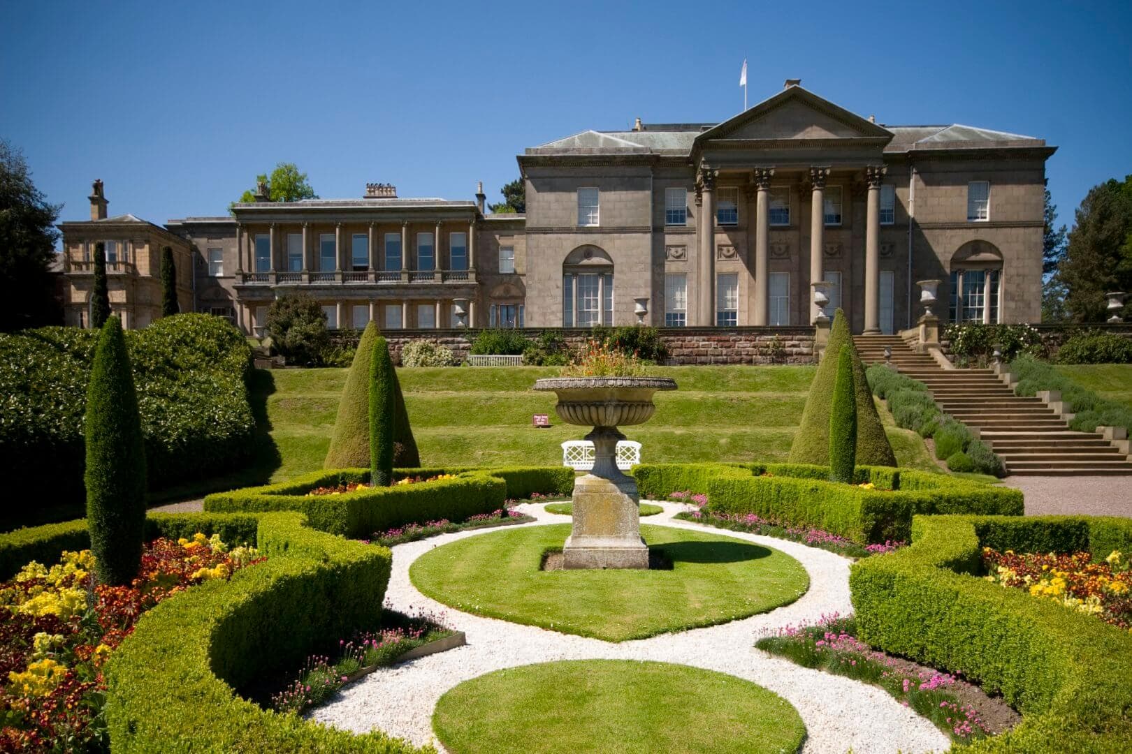 A grand estate with ornate gardens featuring a central stone fountain, manicured hedges, and a neoclassical mansion. - Home Instead