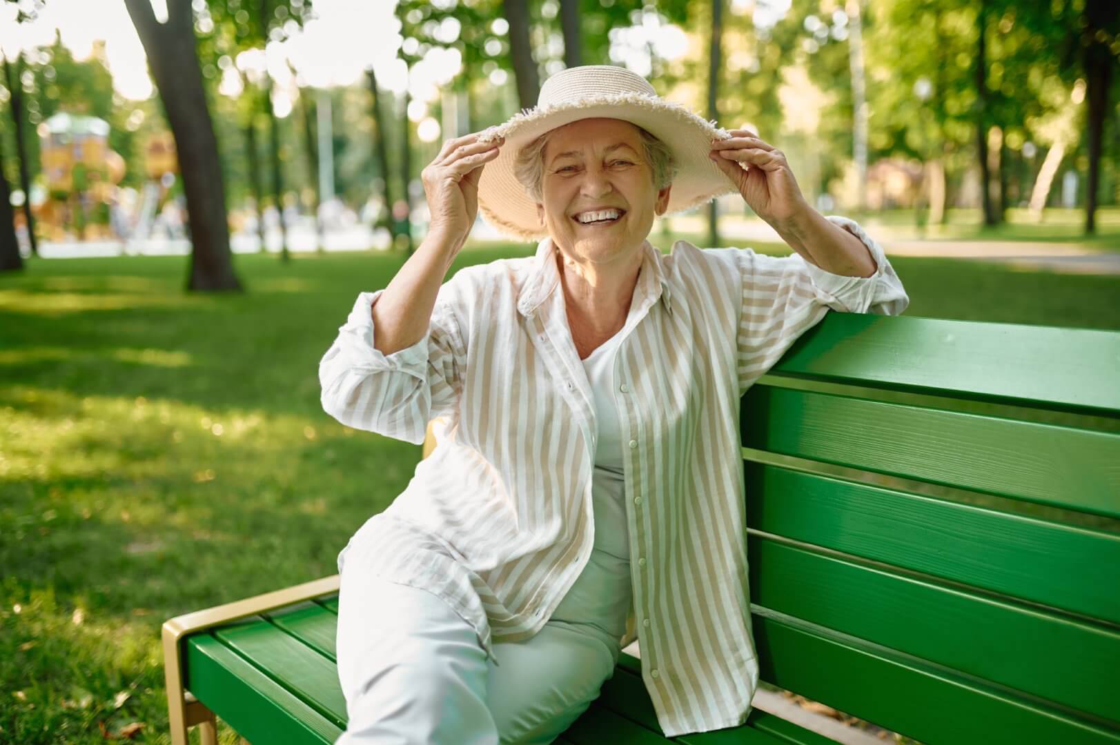 A smiling elderly woman in a hat sits on a green park bench, enjoying a sunny day surrounded by trees. - Home Instead