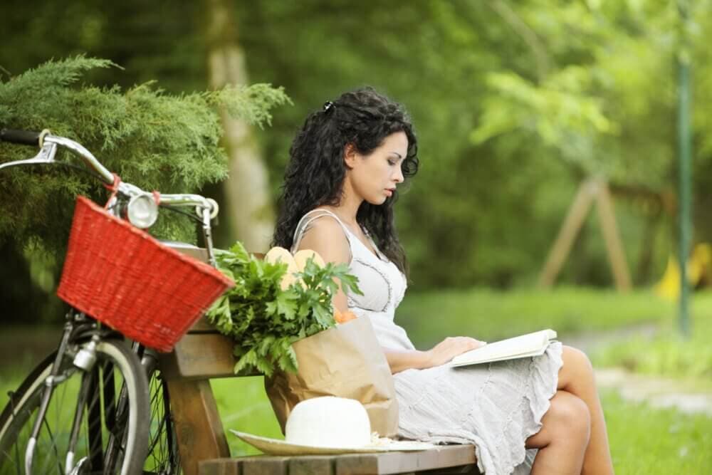 Woman sitting on a park bench reading a book with a bicycle nearby carrying groceries in a basket. - Home Instead