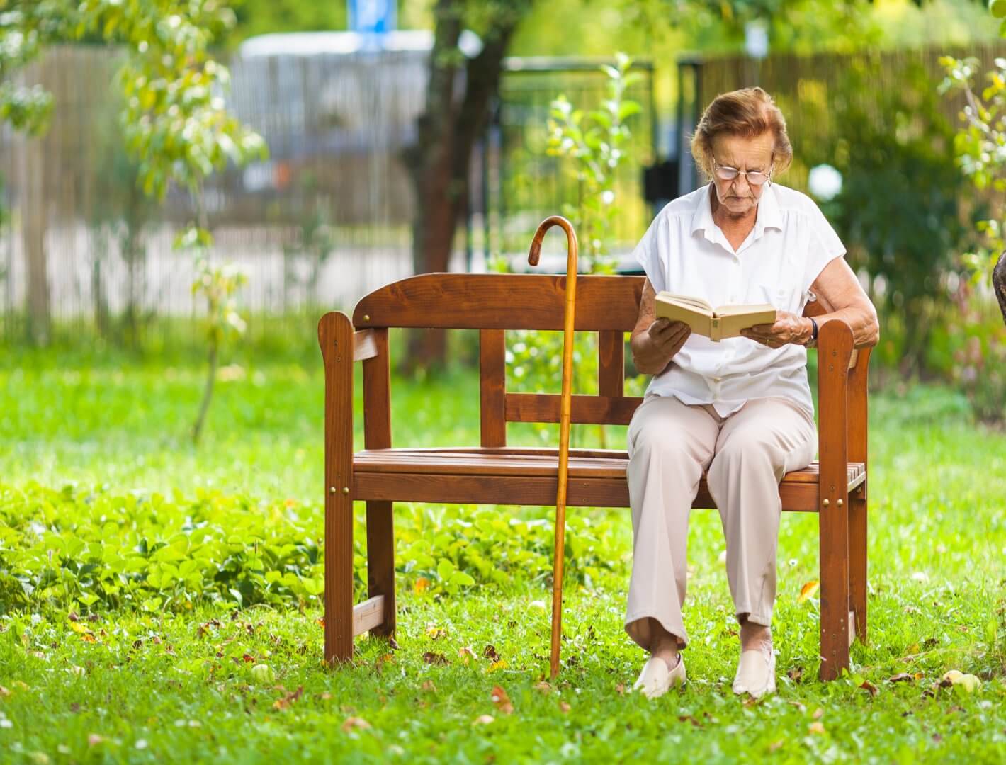 Older woman reading a book on a wooden bench in a garden, with a walking cane resting against the bench. - Home Instead
