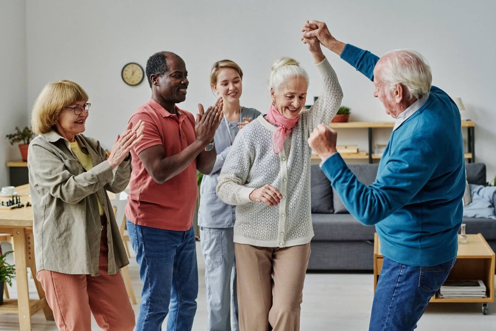 Seniors joyfully dance together in a living room while others clap and smile in the background. - Home Instead
