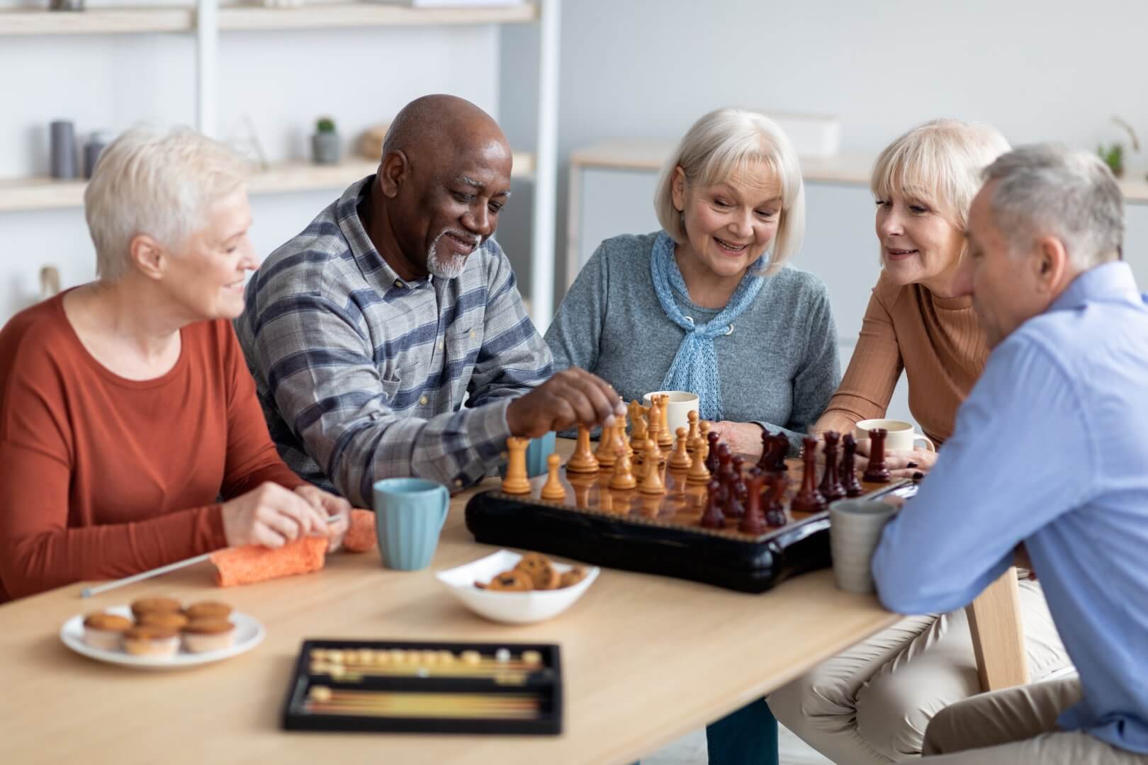 Five elderly people enjoying a game of chess at a table with snacks and drinks, smiling and engaged in conversation. - Home Instead