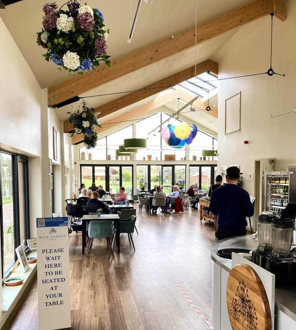 A spacious café with customers seated at tables, greenery, flowers, and bright windows letting in natural light. - Home Instead