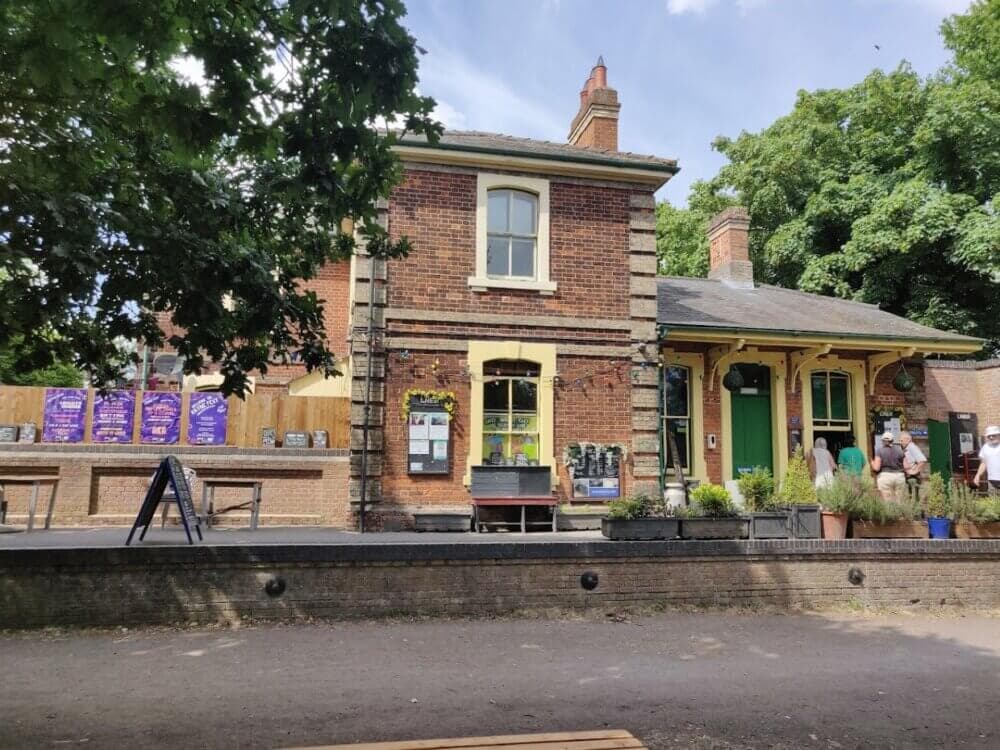 A brick building housing Booking Hall cafe Rayne with green and yellow trim, surrounded by trees and people standing outside. - Home Instead