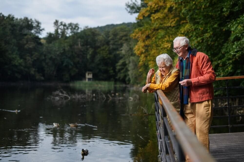An elderly couple smiling and leaning on a railing while looking at a pond in a park with trees in the background. - Home Instead
