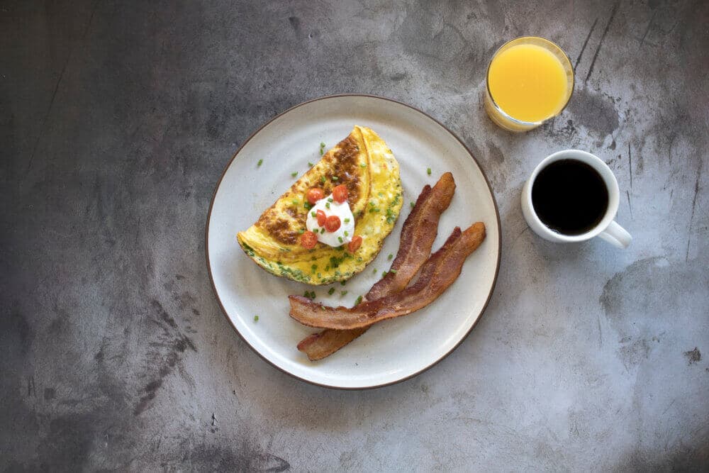 A plate with an omelet, bacon strips, a mug of coffee, and a glass of orange juice on a textured surface. - Home Instead