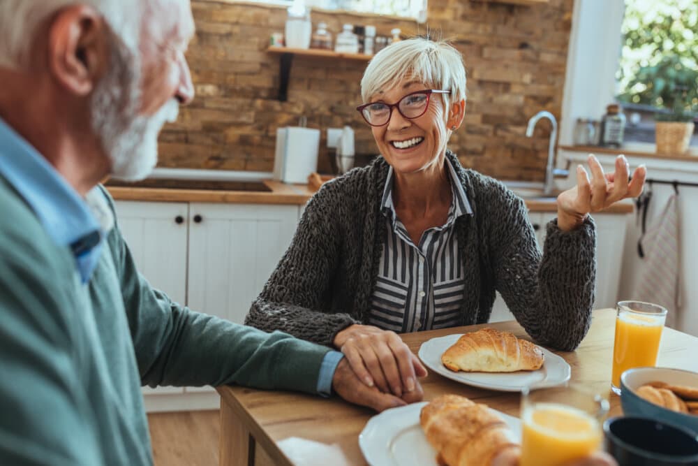 Elderly couple enjoying breakfast at a kitchen table, smiling and talking. Croissants and orange juice are on the table. - Home Instead