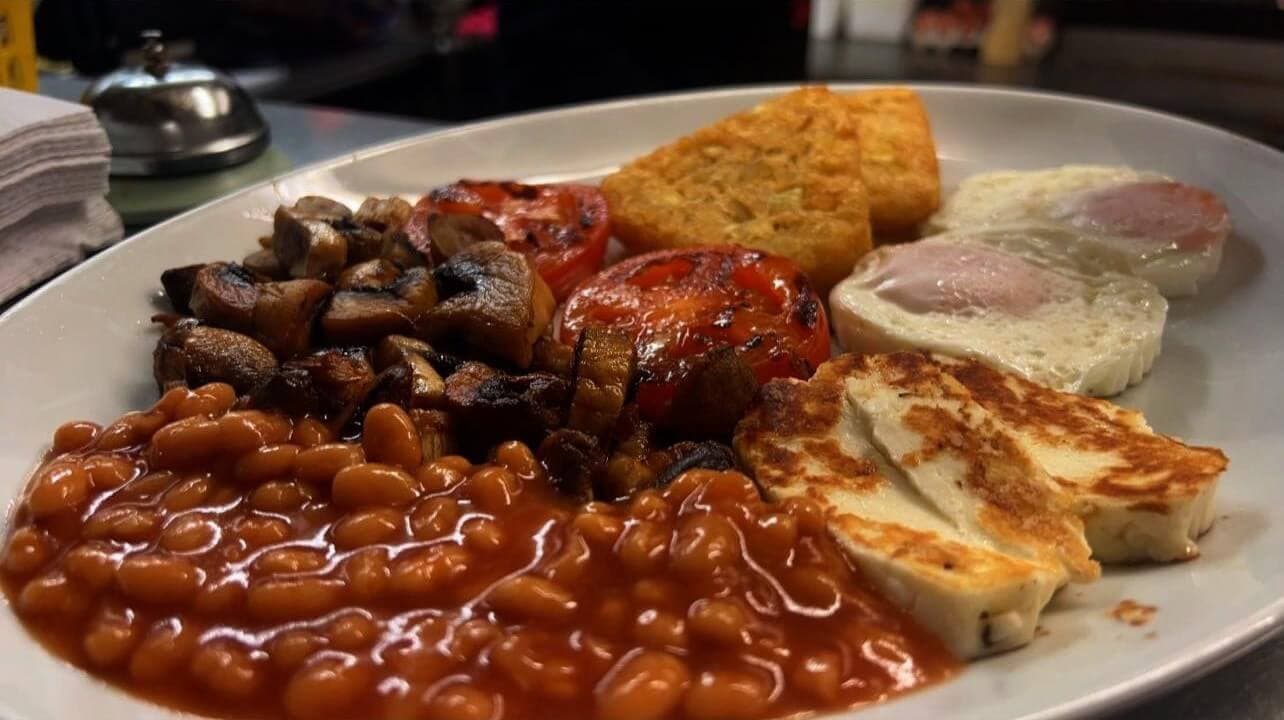 A plate of beans, mushrooms, grilled tomatoes, hash browns, fried eggs, and cheese on a table. - Home Instead