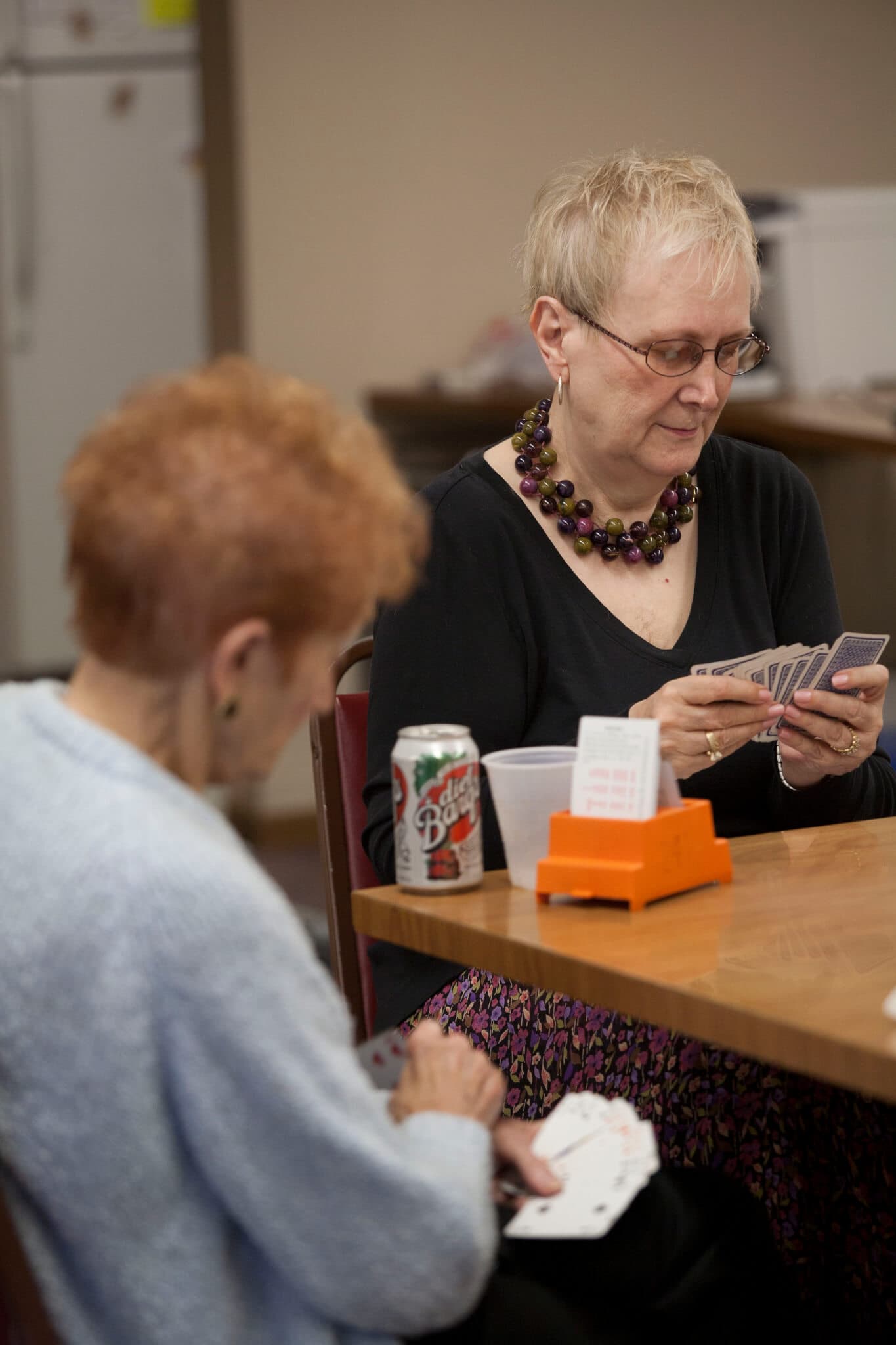Two elderly women engaged in a card game at a table, one woman has a can of Dr Pepper next to her. - Home Instead