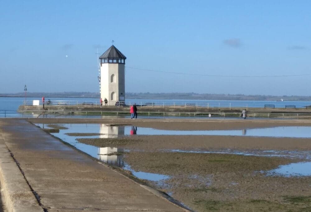 Lighthouse on a pier with its reflection in water, under a clear blue sky. Two people walk nearby. - Home Instead