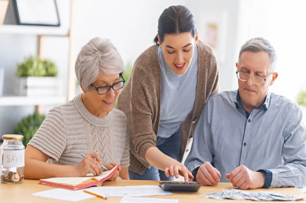 A young woman helps an older couple with financial planning, using a calculator and notebook, at a table. - Home Instead