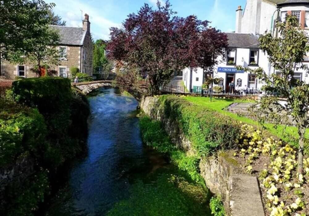 A scenic view of a clear stream flowing by stone buildings and lush greenery under a sunny, blue sky. - Home Instead