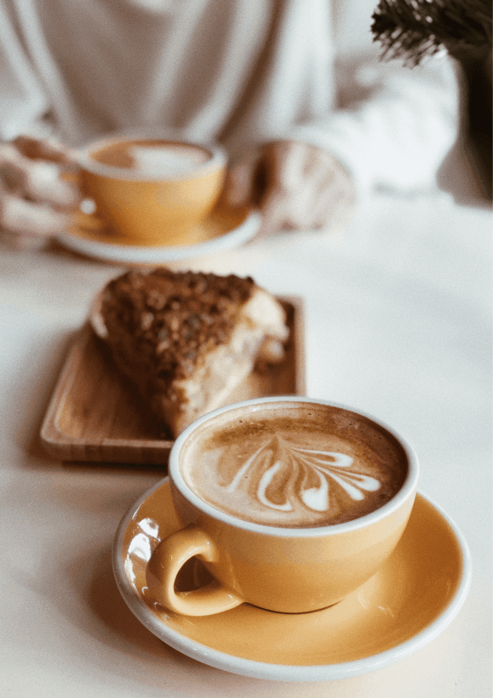 Two cups of coffee in yellow mugs on a table with a slice of cake. A person reaches for a cup in the background. - Home Instead