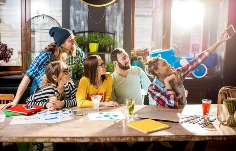 A group of five people sitting at a table with work materials, posing for a selfie in a bright, casual setting. - Home Instead