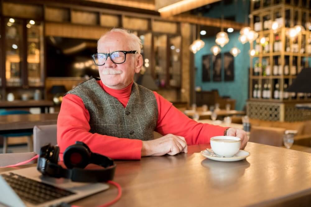 Older man in glasses, red shirt, and grey vest sits at a café table with coffee, laptop, and headphones. - Home Instead Southampton