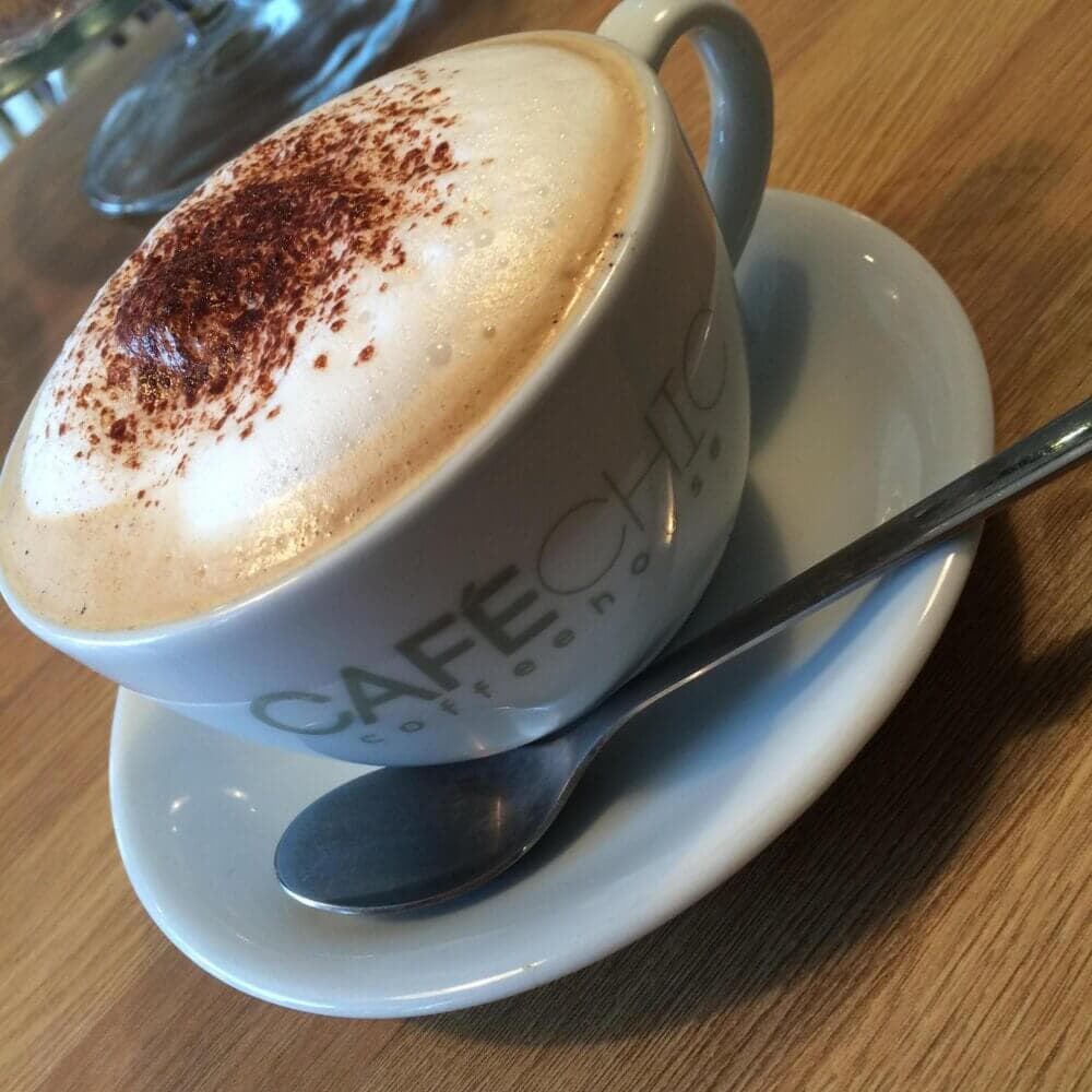 A cup of cappuccino with foam and cocoa powder on top, placed on a saucer with a spoon, on a wooden table. - Home Instead