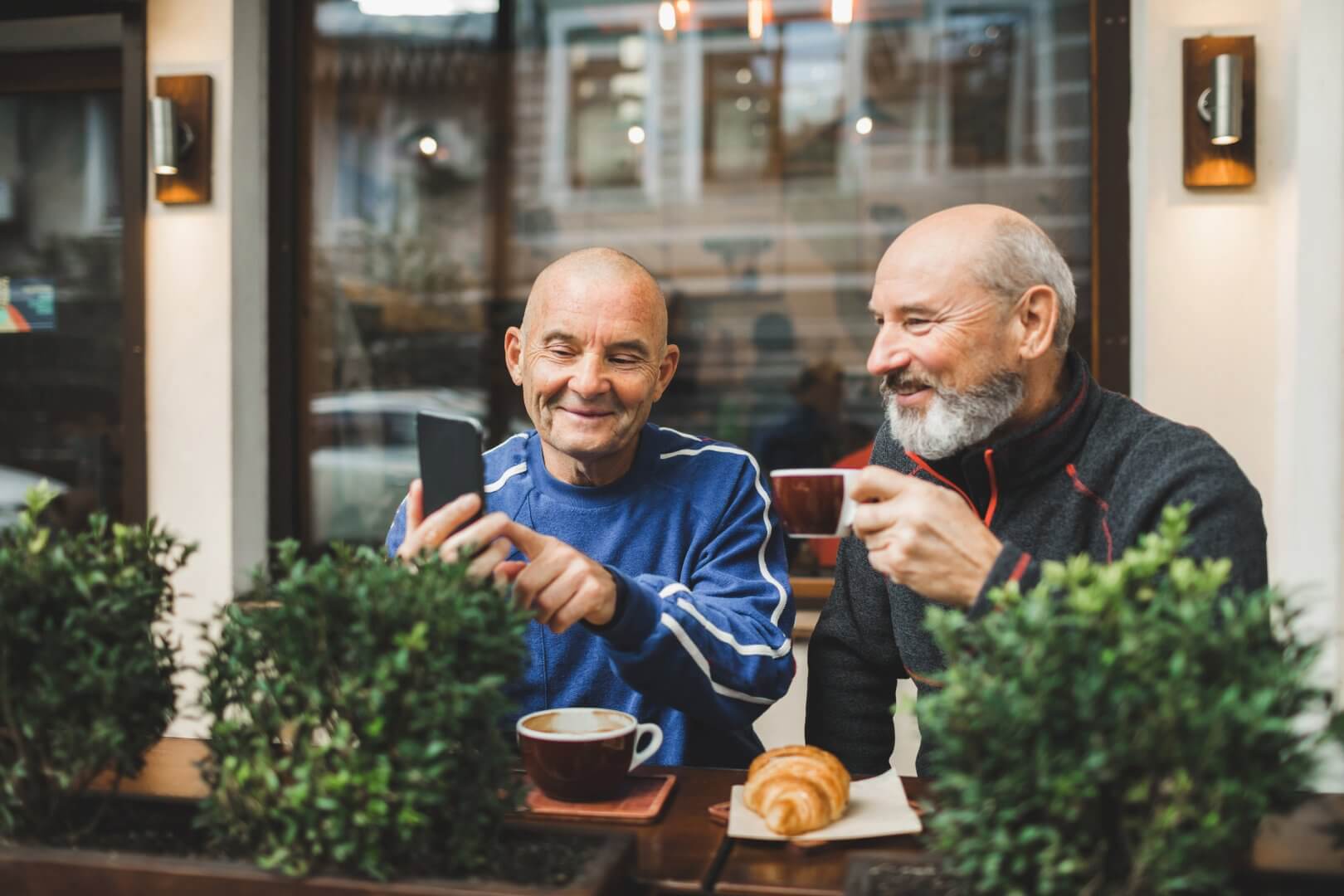 Two older men enjoying coffee and pastries while taking a selfie at an outdoor cafe. - Home Instead