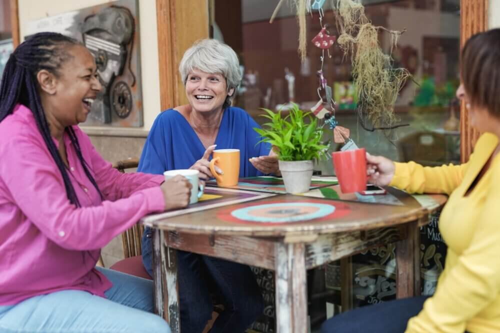 Three women sitting at a rustic table, laughing and drinking coffee together in an outdoor cafe setting. - Home Instead