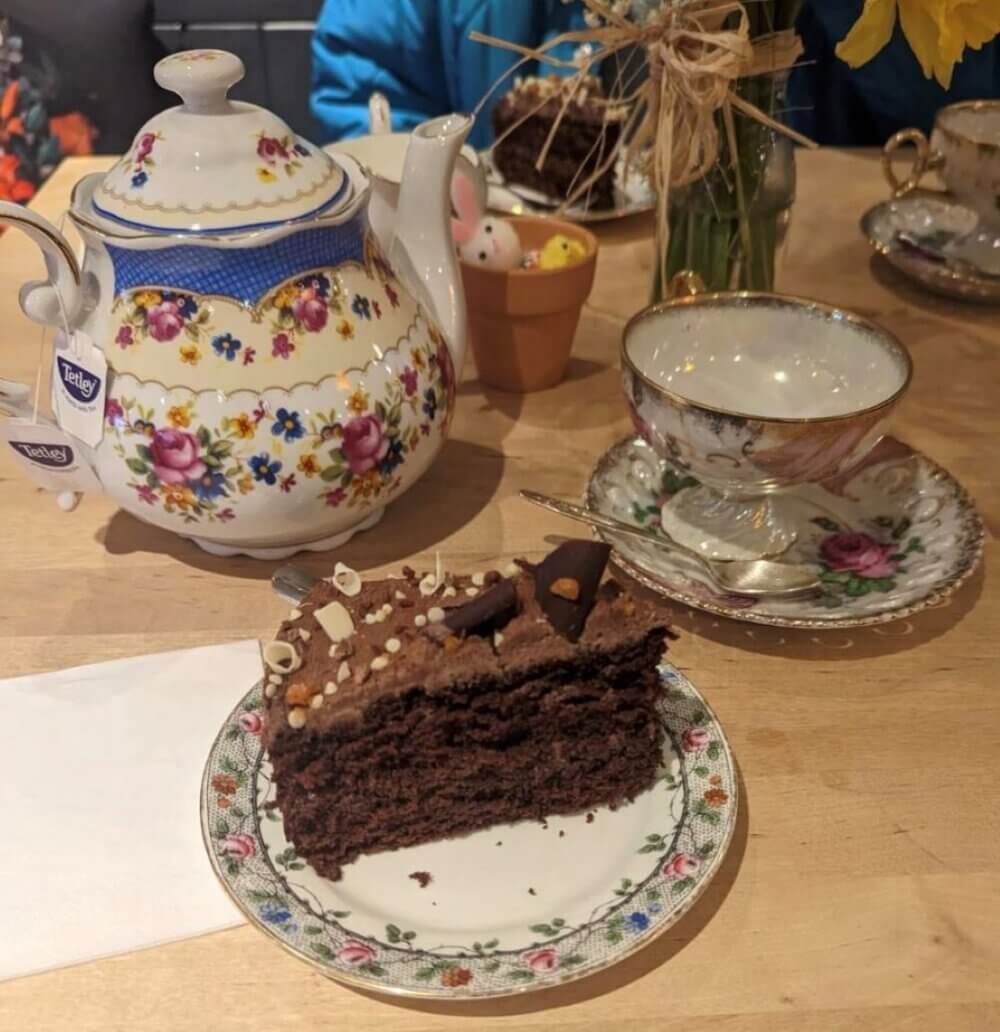 A slice of chocolate cake on a floral plate, next to a teapot and teacup on a wooden table. - Home Instead