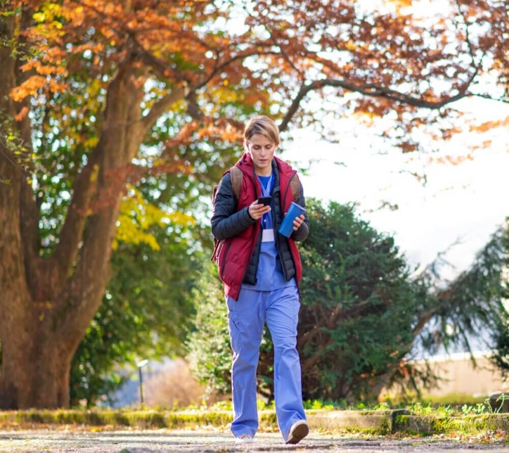 Person wearing a red vest and blue scrubs walking outdoors while looking at their phone during a sunny autumn day. - Home Instead