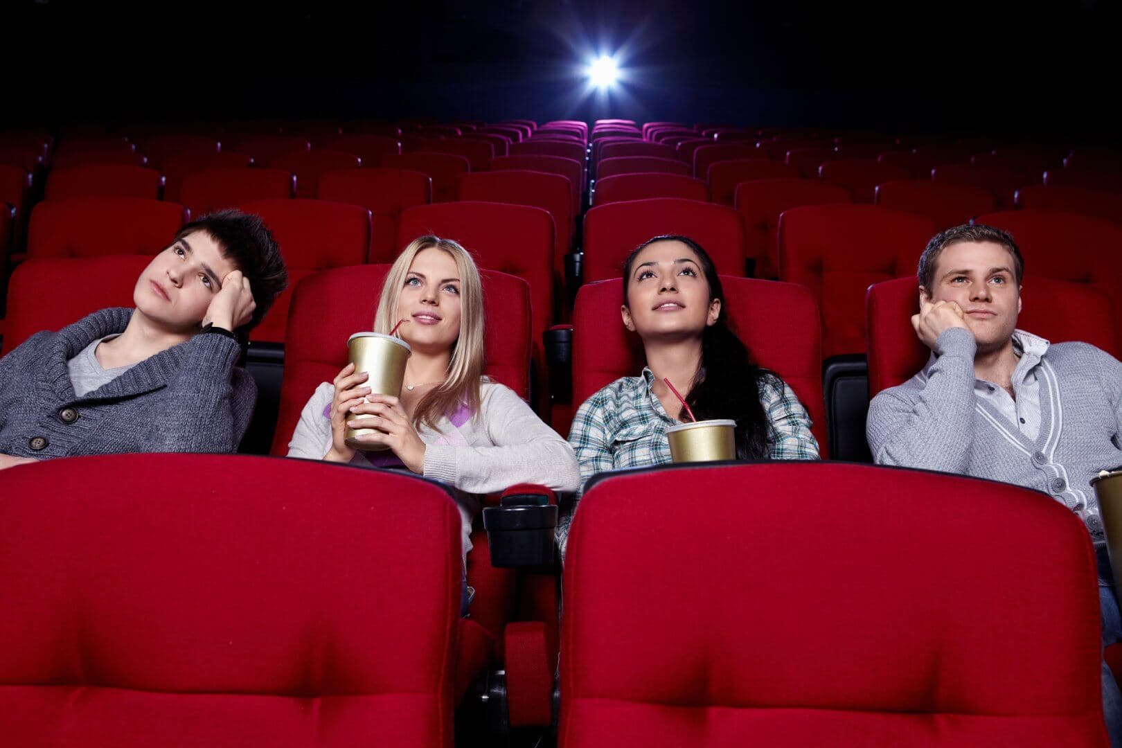 Four people sitting in red theater seats, watching a movie. They are holding drinks and appear engaged with the screen. - Home Instead