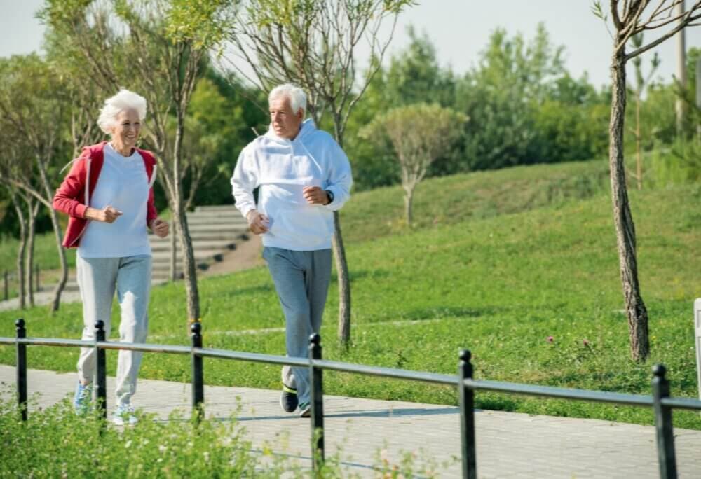 Elderly couple jogging on a paved path through a park, surrounded by greenery and trees. - Home Instead