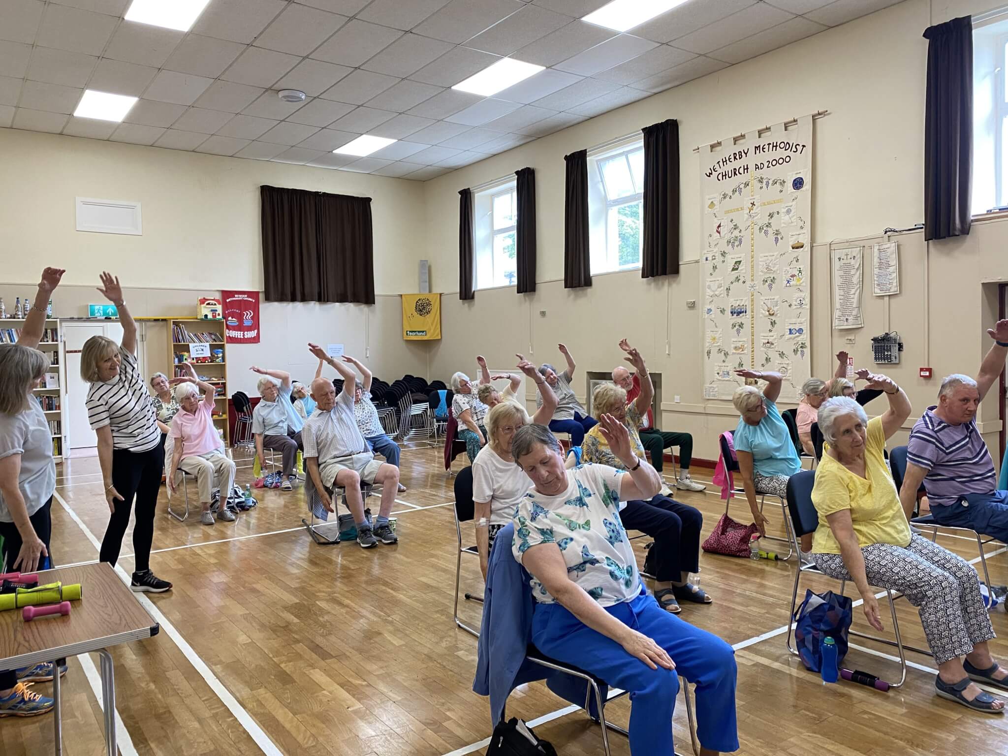 A group of seniors participate in a seated exercise class in a community hall, raising one arm in unison. - Home Instead