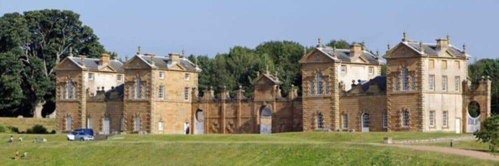 A historic estate with symmetrical stone buildings, archways, and surrounded by greenery under a bright blue sky. - Home Instead